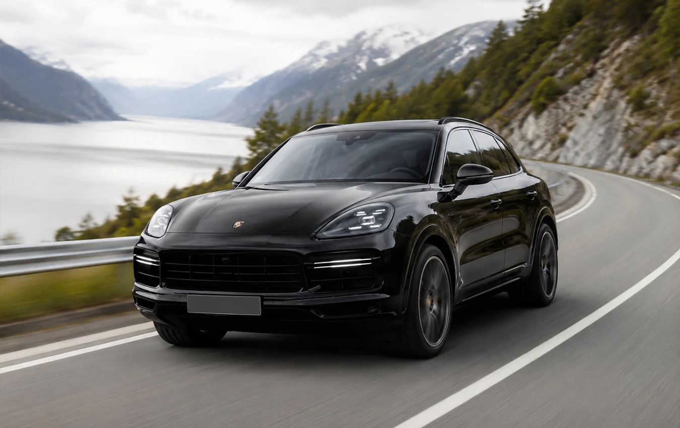 Schwarzer Porsche Cayenne SUV fährt auf einer kurvigen Küstenstraße vor einer Berg- und Seekulisse.